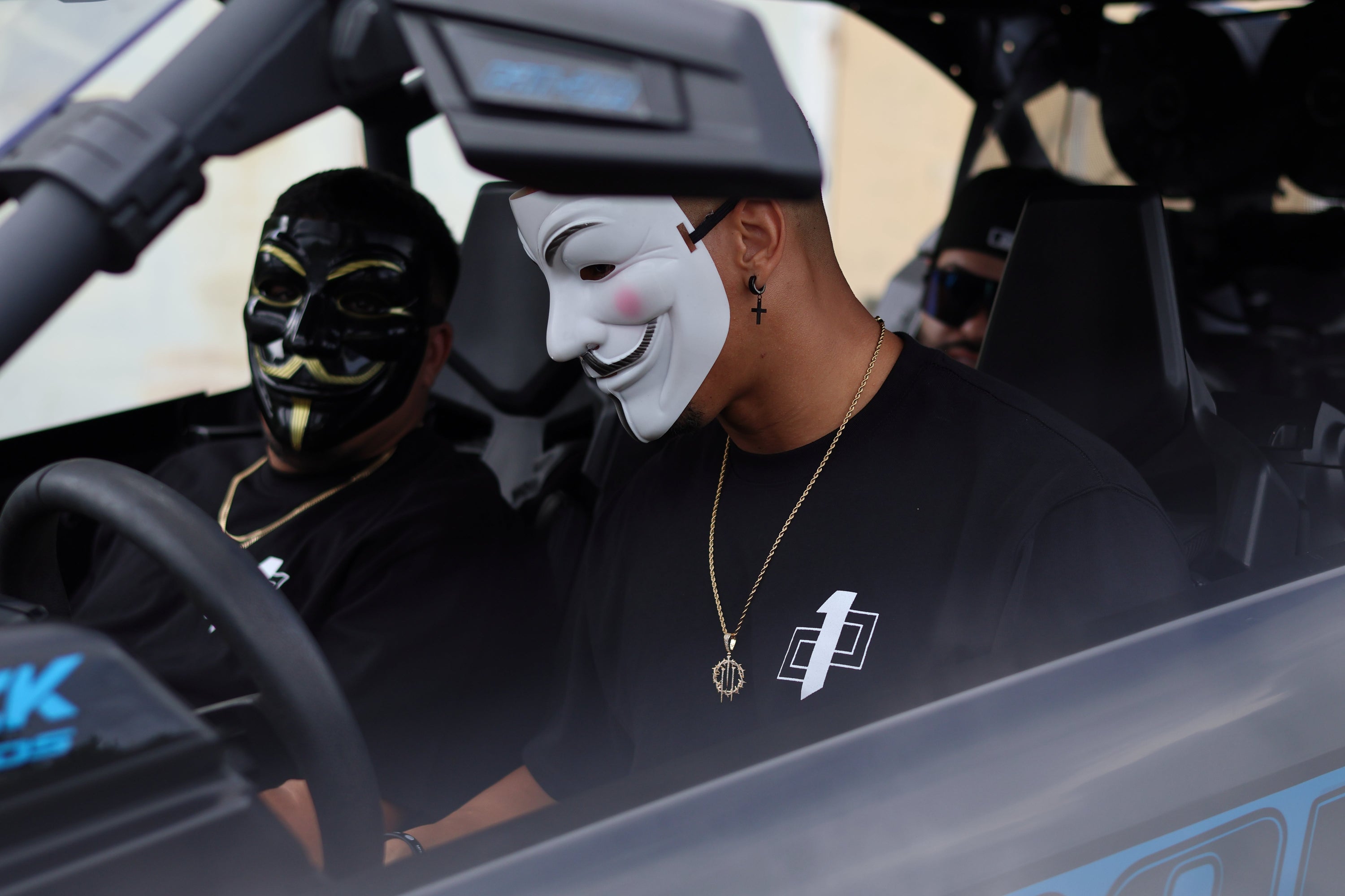 Two individuals wearing masks sitting in a car with one shirt displaying the 1 percent mentality symbol